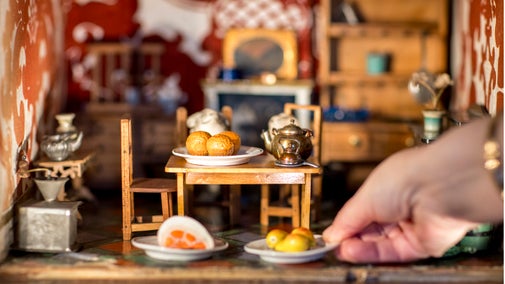 Food and a teapot are on a table in a tiny dolls house room. A hand places a tiny plate of pears on the floor in front of the table.